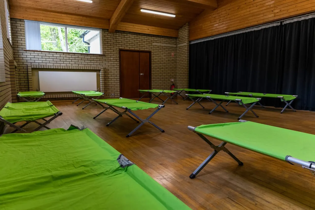 Indoor hall with multiple bright green camp beds arranged on wooden flooring, wooden ceiling and brick walls.