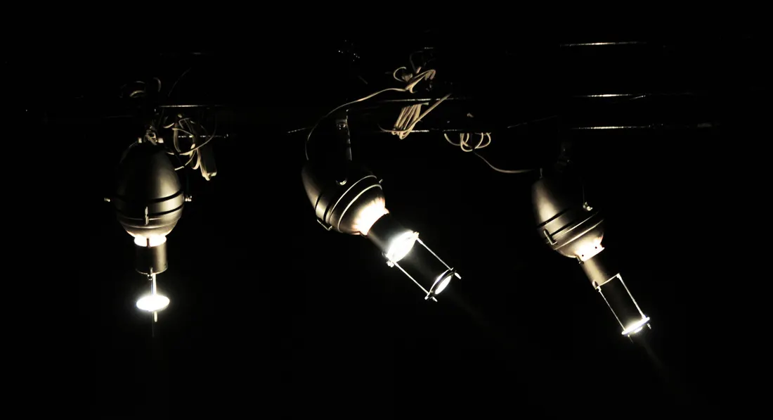 Three stage lights suspended against a black background, illuminated and casting downward beams.