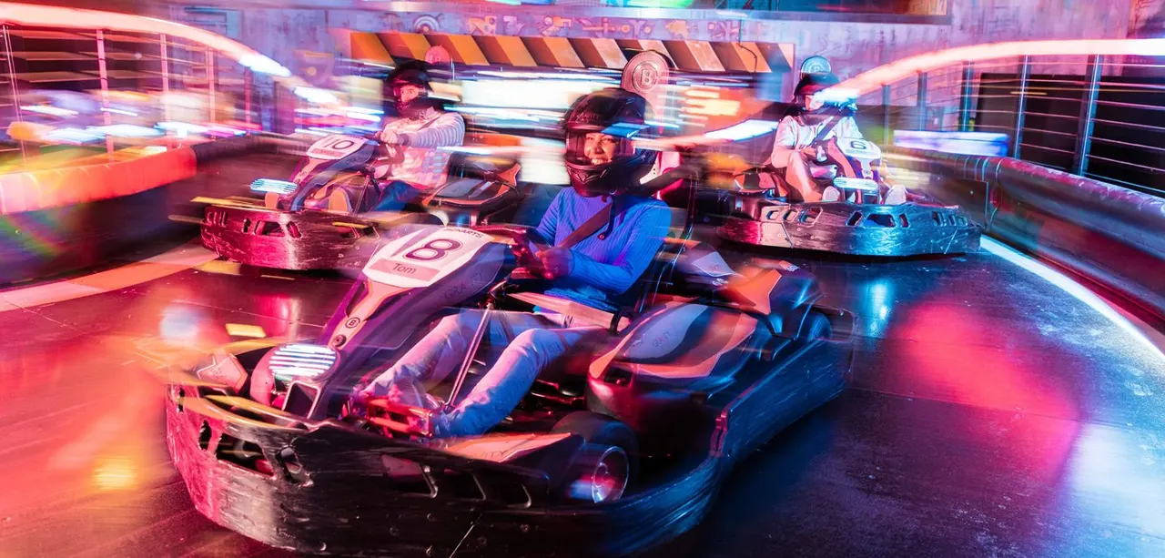 Racers in electric go-karts on an indoor track lit with vibrant neon pink and blue lighting.