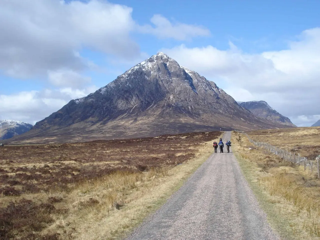 Hikers walking along a remote gravel track towards a snow-capped mountain in a barren highland landscape.