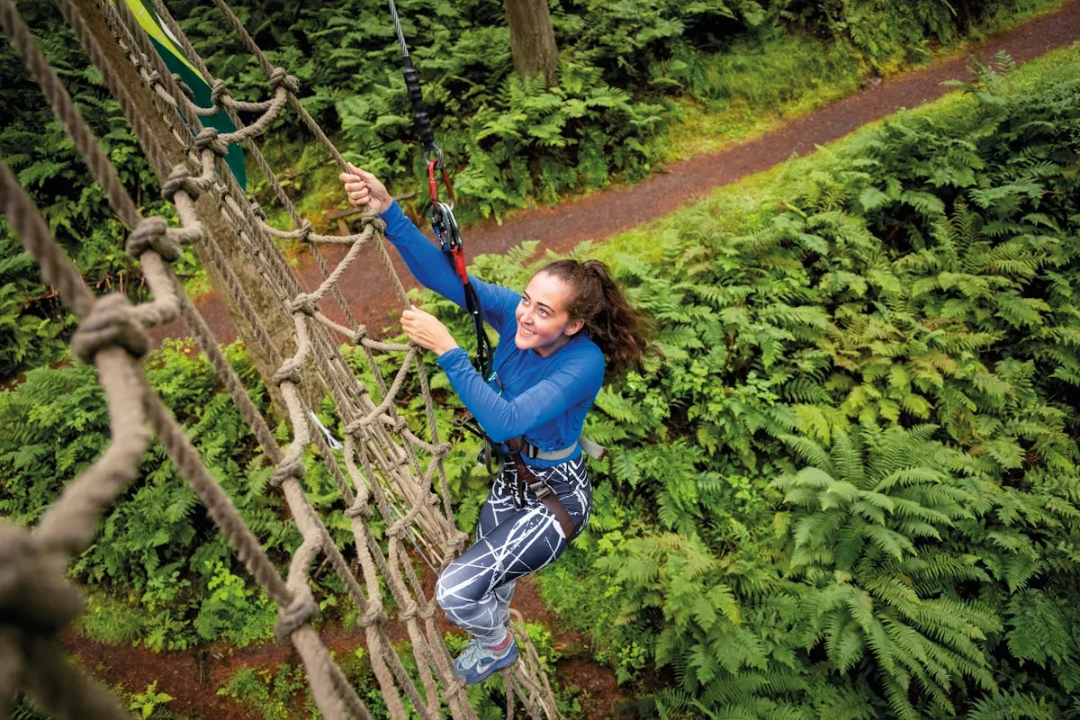 Young woman climbing rope obstacle course above forest canopy surrounded by green trees.