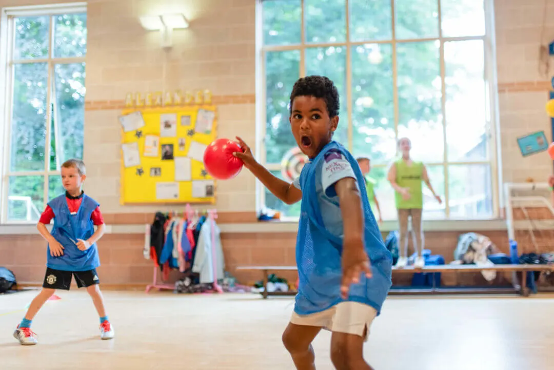 Children playing dodgeball indoors in a school gymnasium with large windows and a yellow noticeboard visible.