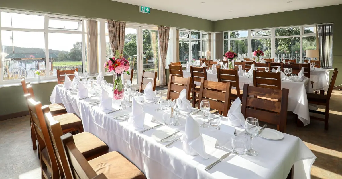 Bright dining room with long white-clothed tables set for a formal meal, wooden chairs, pink floral centrepieces, and countryside views through large windows.