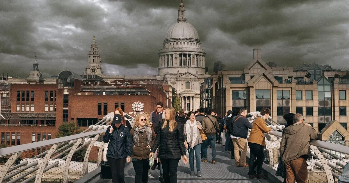 Visitors walking across a pedestrian bridge with St Paul's Cathedral visible in the background under dramatic cloudy skies.