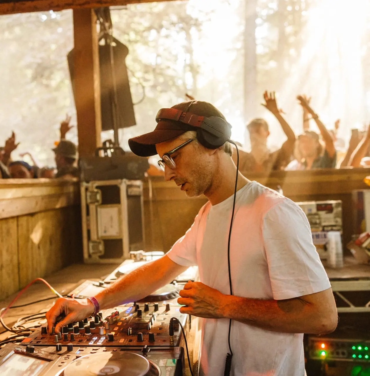 DJ operating mixing equipment at an outdoor festival with dancing crowd in background.