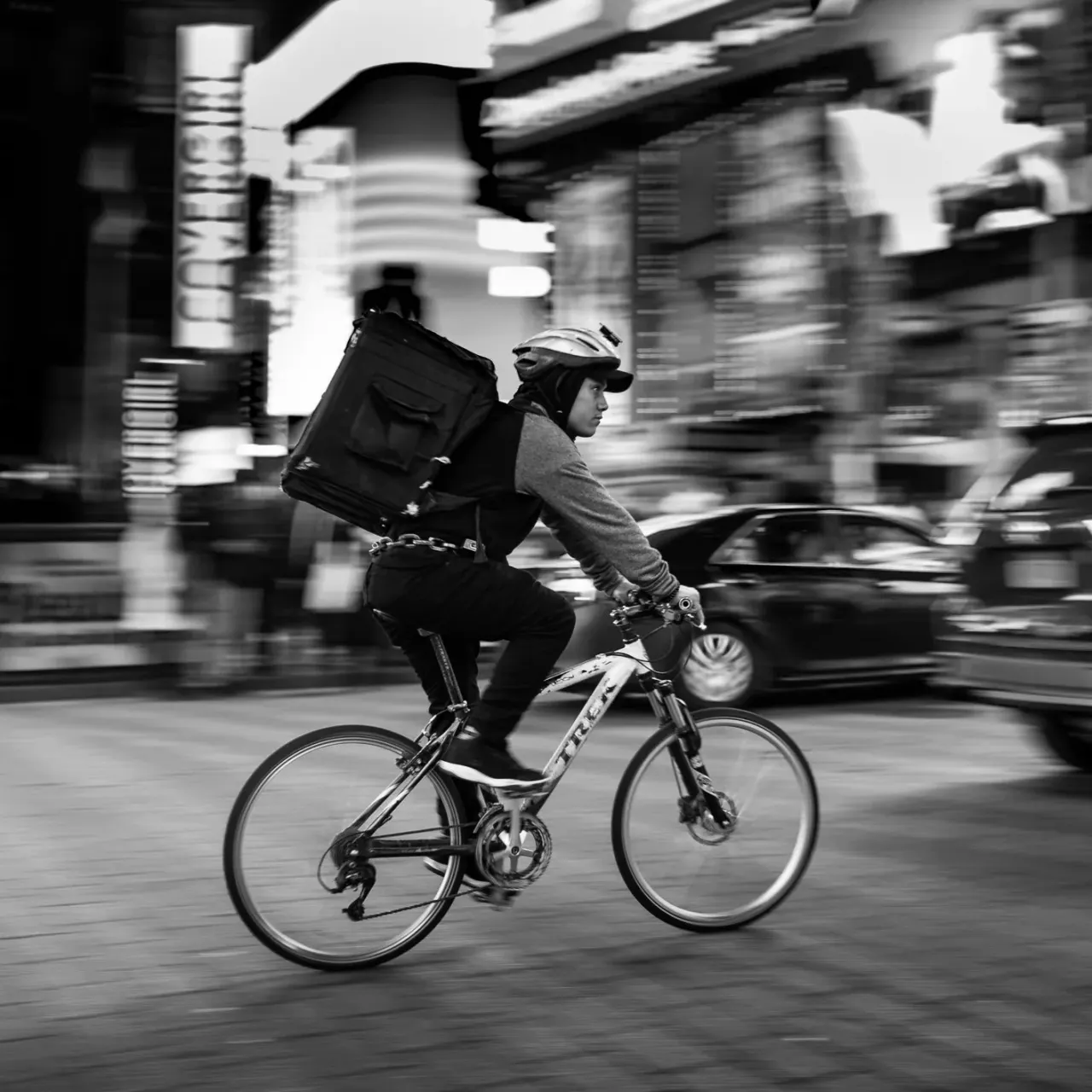 Person cycling through a city street wearing a helmet and delivery backpack.