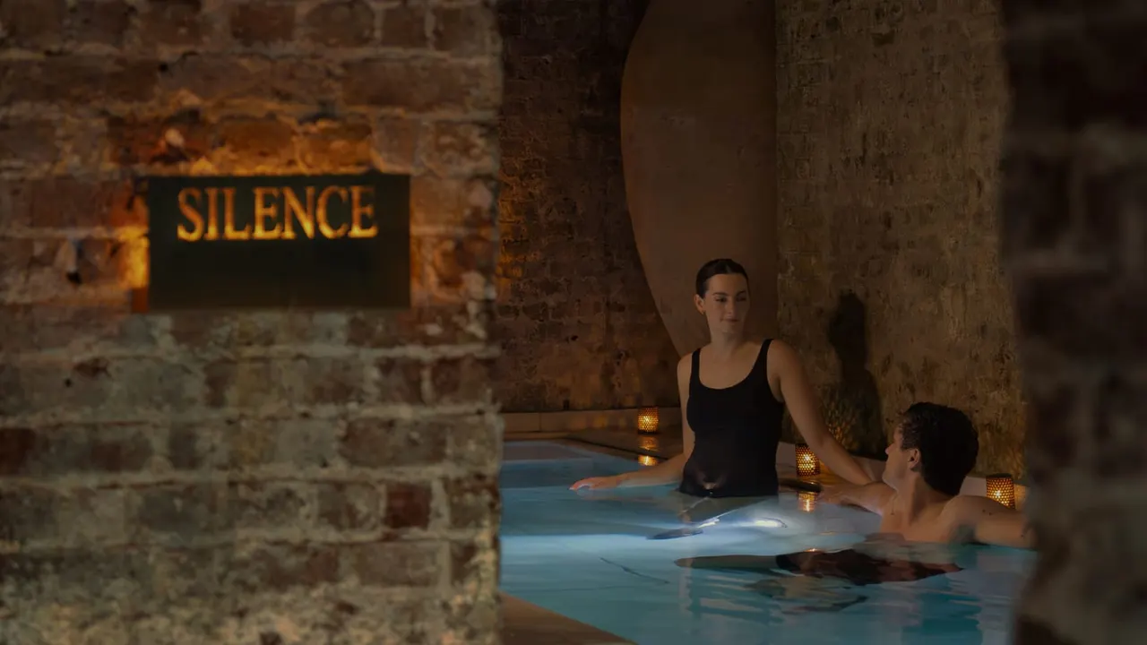 Two people bathing in a thermal pool within a stone-vaulted medieval chamber lit by candlelight.