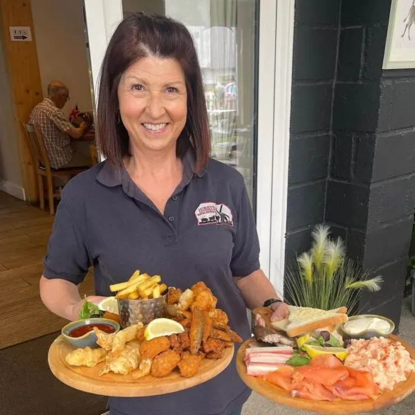 Staff member holding wooden boards displaying fish and chips, seafood platters, and chips at a farm shop cafe.
