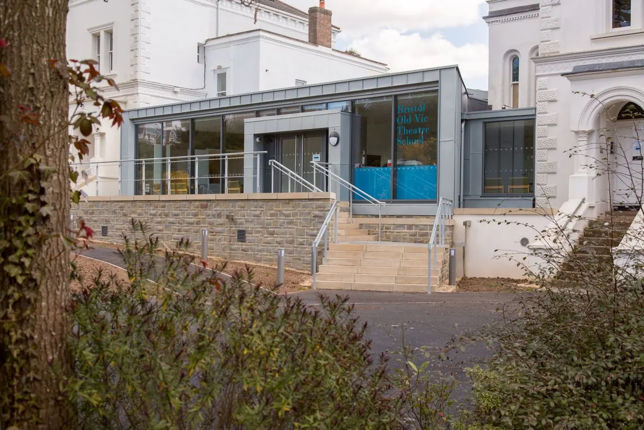 Modern glass and steel extension to a historic white stone building, with metal railings and steps leading to the entrance of Bristol Old Vic Theatre School.
