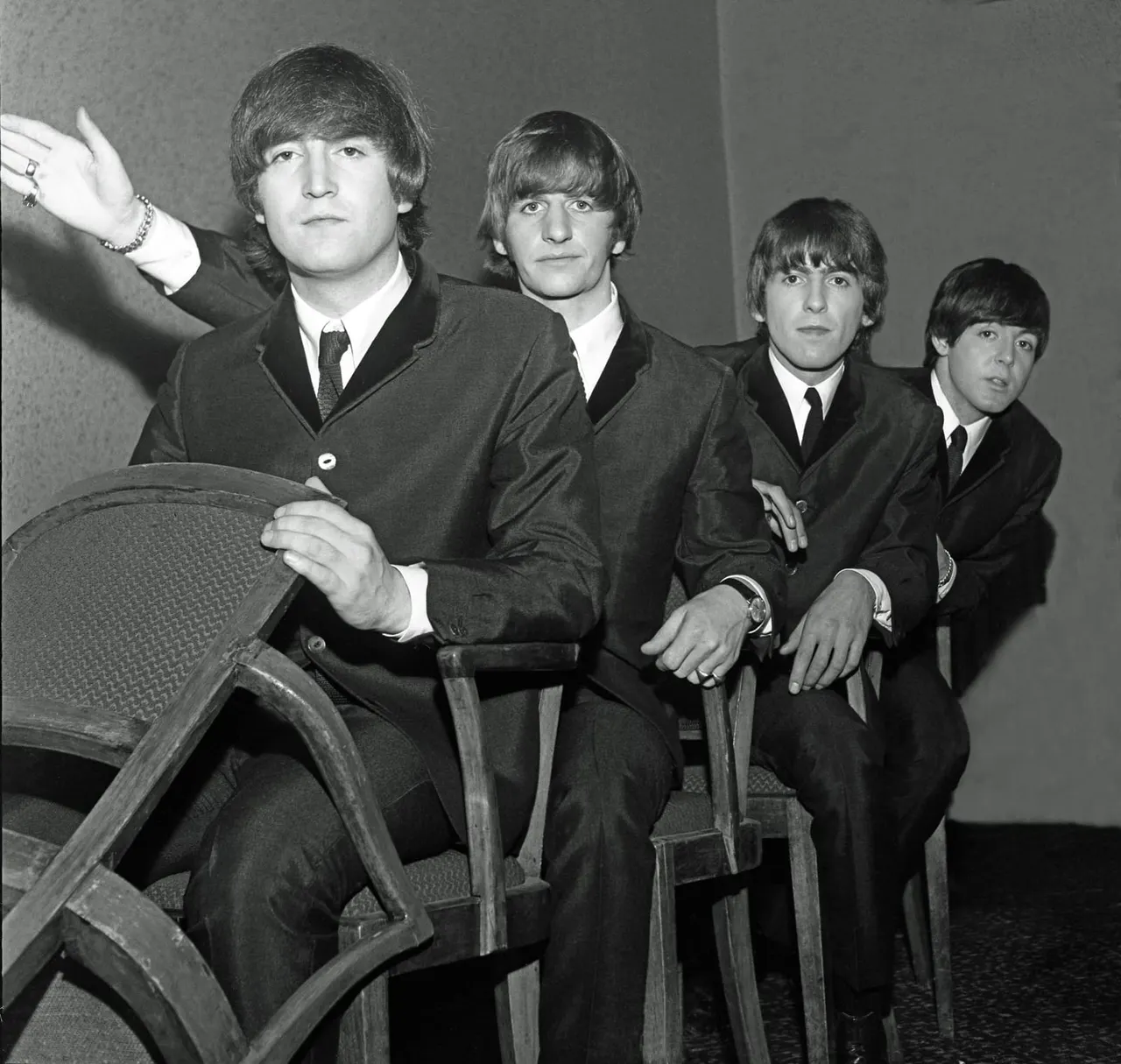 Five men in 1960s suits and matching haircuts seated on chairs in black and white photograph.