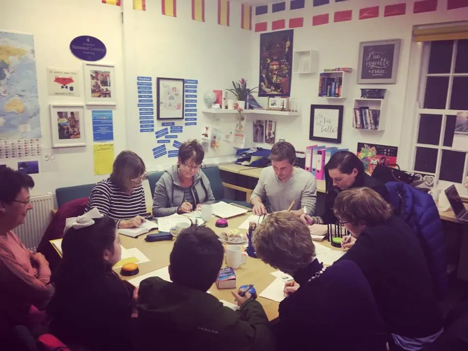 A group of adults seated around a table in a classroom, engaged in a language learning session with notebooks and refreshments.