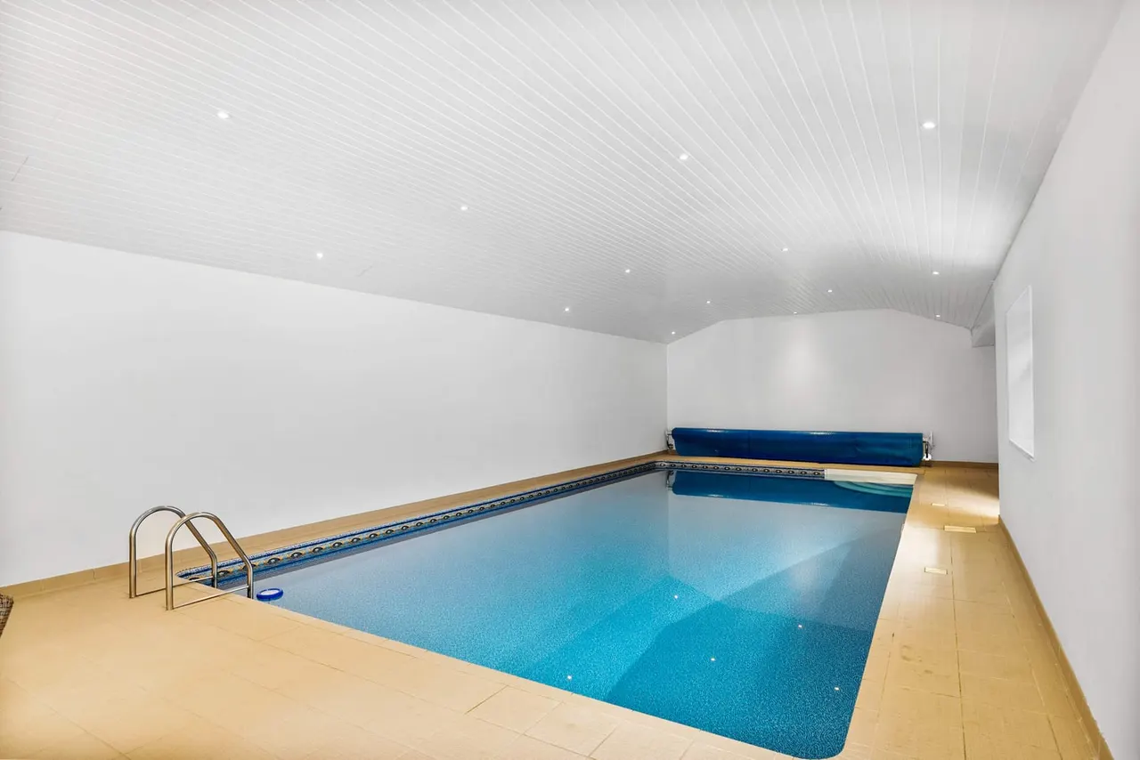 Indoor swimming pool with turquoise water, metal ladder and blue cover, in a white-walled room with wooden decking.