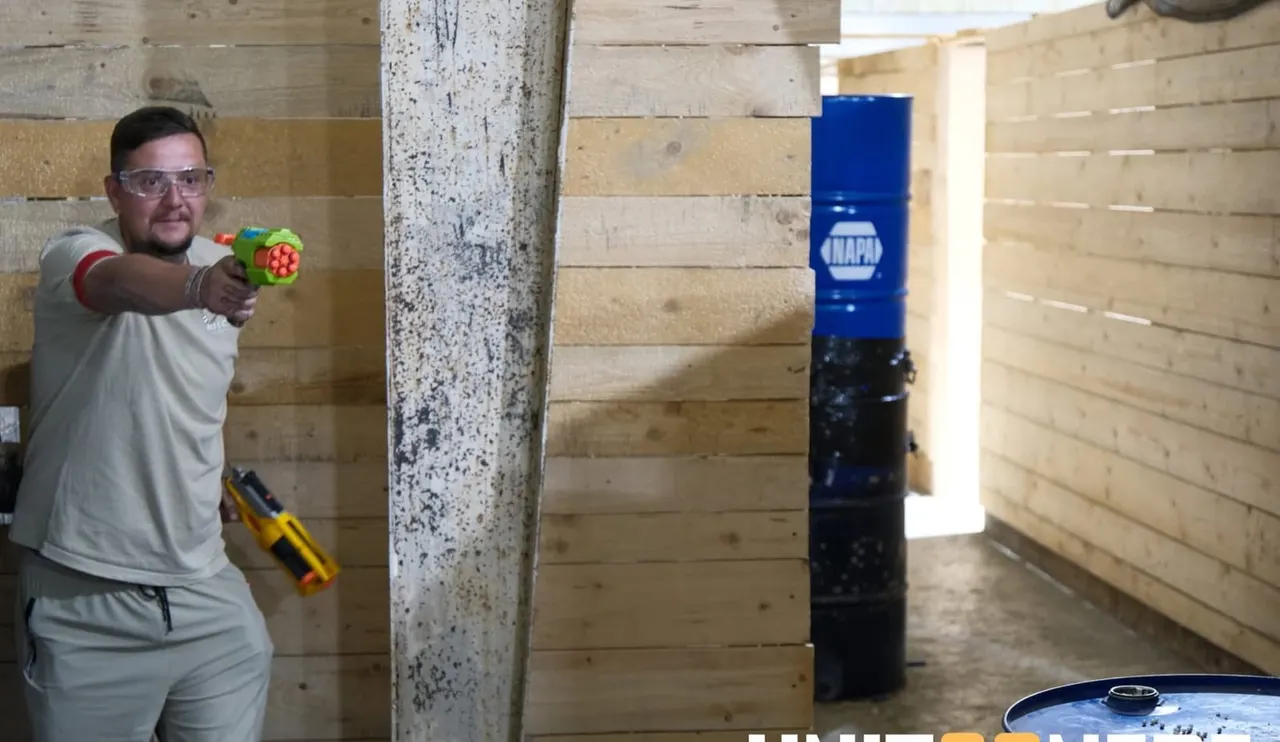 Man in safety glasses aiming foam dart blaster at target in wooden-walled indoor airsoft arena.