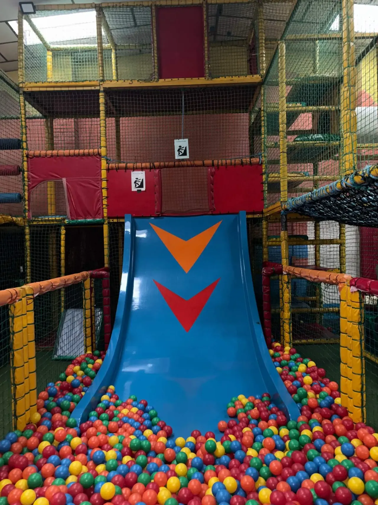 Blue slide with orange and red chevron design descending into coloured ball pit inside indoor play structure.