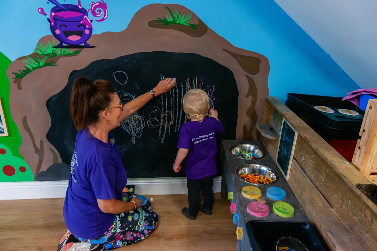 An adult and young child drawing on a large blackboard cave structure during a creative activity session.