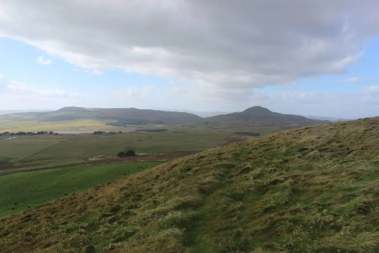 Rolling hillside with grassland and heather overlooking a distant valley with two volcanic peaks beneath cloudy sky.