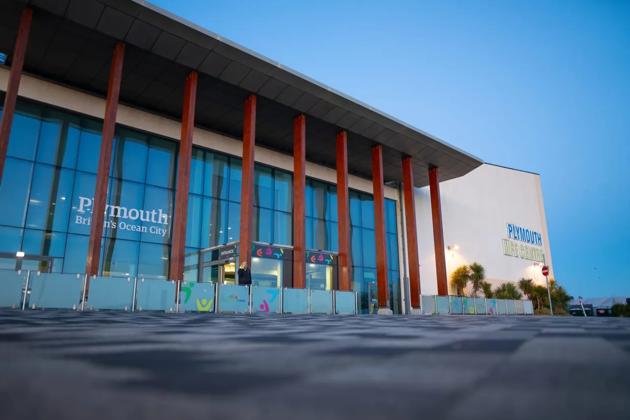Modern leisure centre building with red pillars, glass frontage and palm trees against clear blue sky.