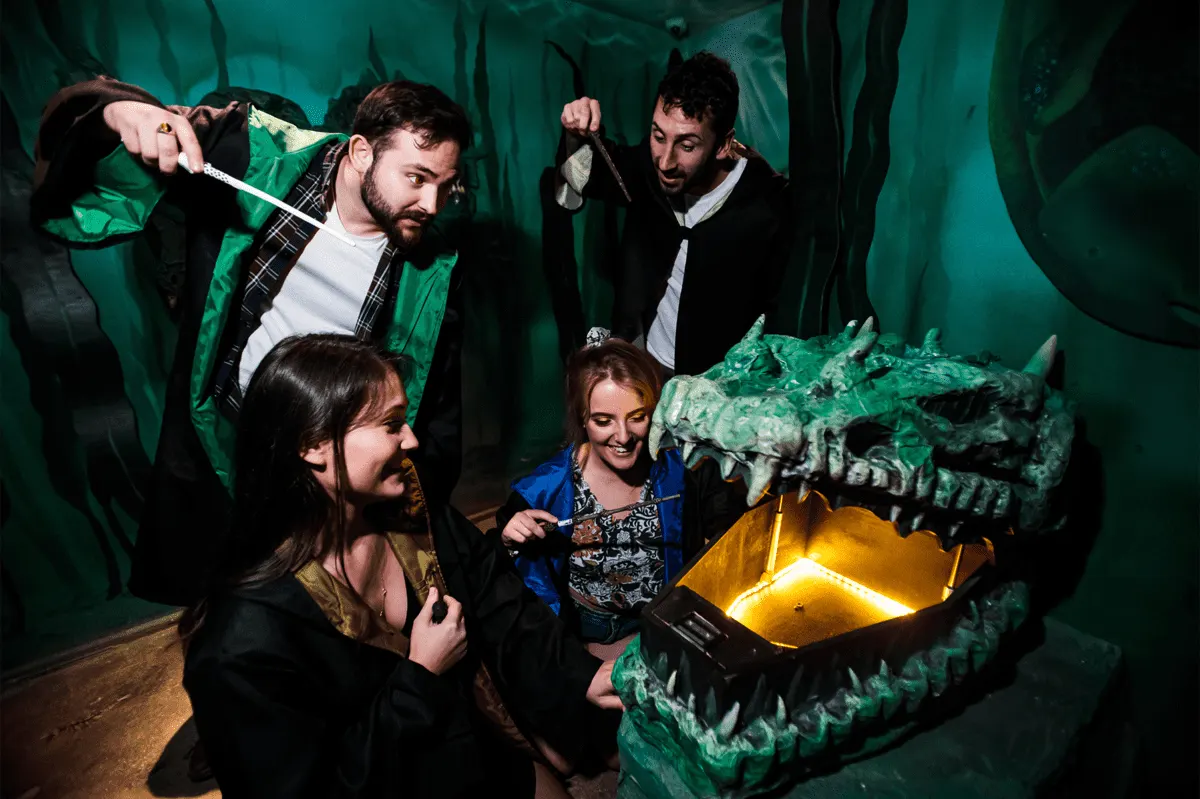 Four people inside a dragon-themed escape room with glowing eyes and mouths, holding torches and examining props.