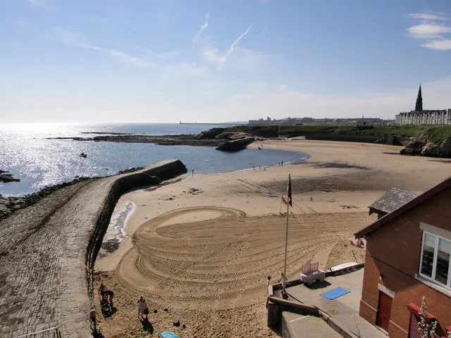 Sandy beach with visitors, rock groyne, church spire on horizon, and calm blue sea on a sunny day.