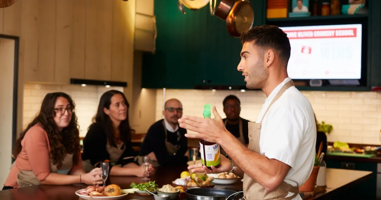 Man in white shirt conducting a cooking class demonstration to seated guests around a kitchen counter with food and ingredients.