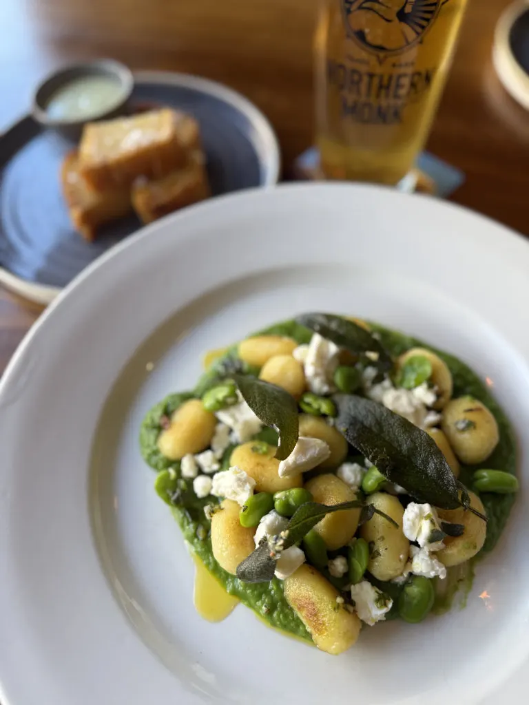 White bowl of spring vegetables with pea purée, topped with cheese and sage leaves, served with bread and beer.