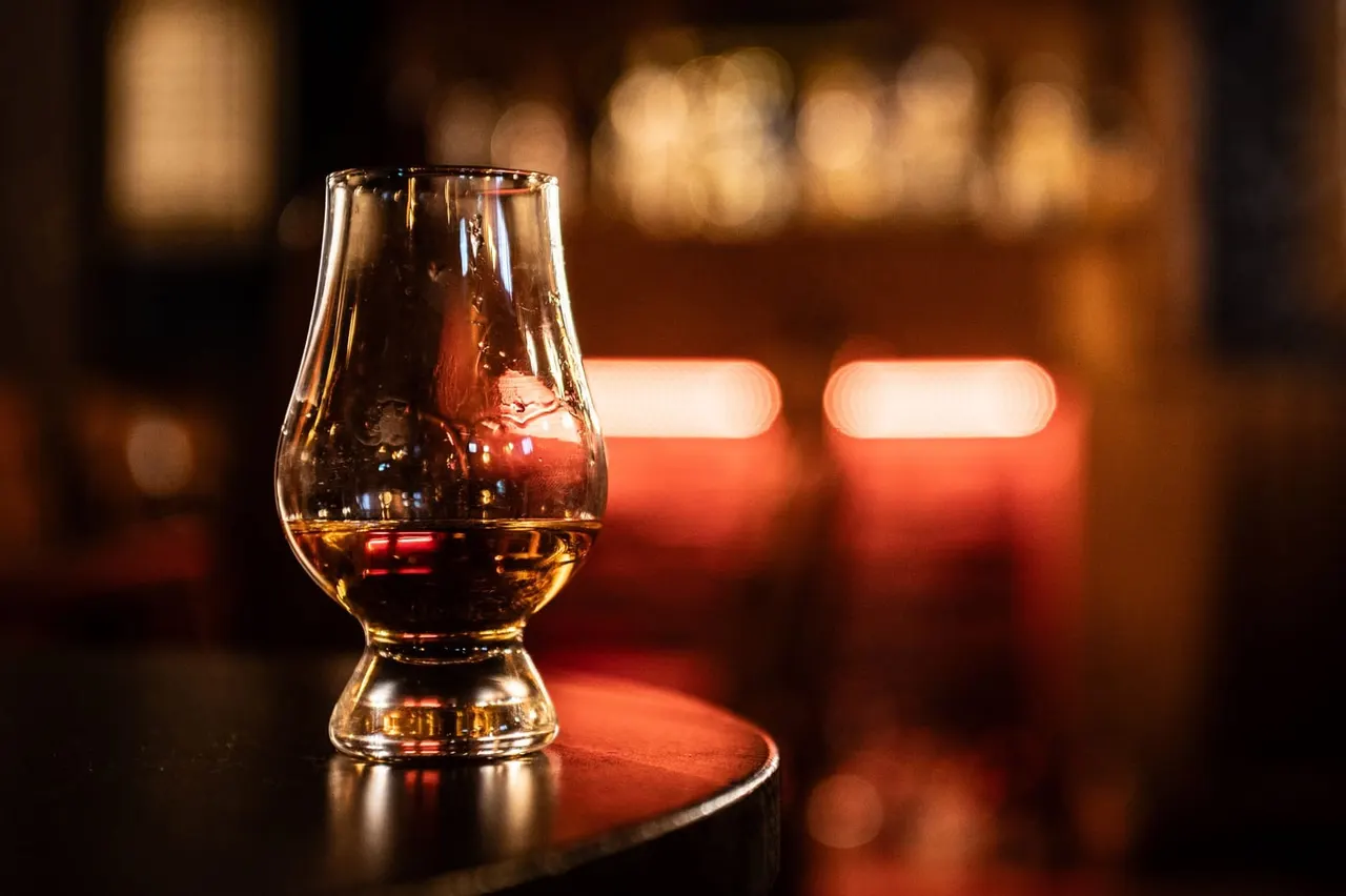 A glass of whisky on a dark table with warm, blurred lighting in the background.