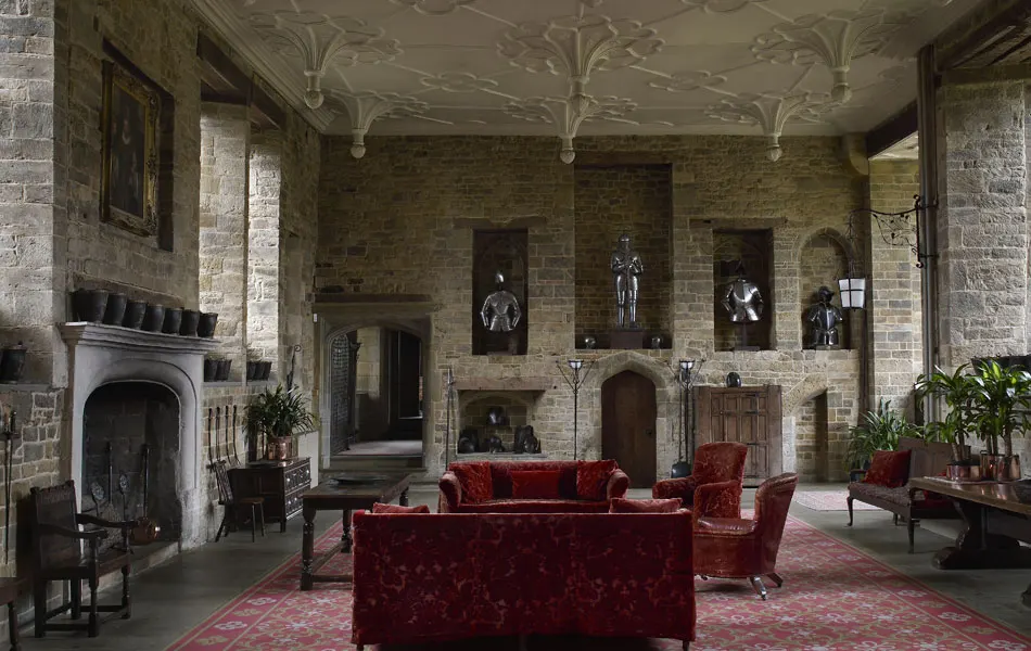 Grand stone hall with ornate ceiling, red velvet furniture, fireplace, and artwork displayed on brick walls.
