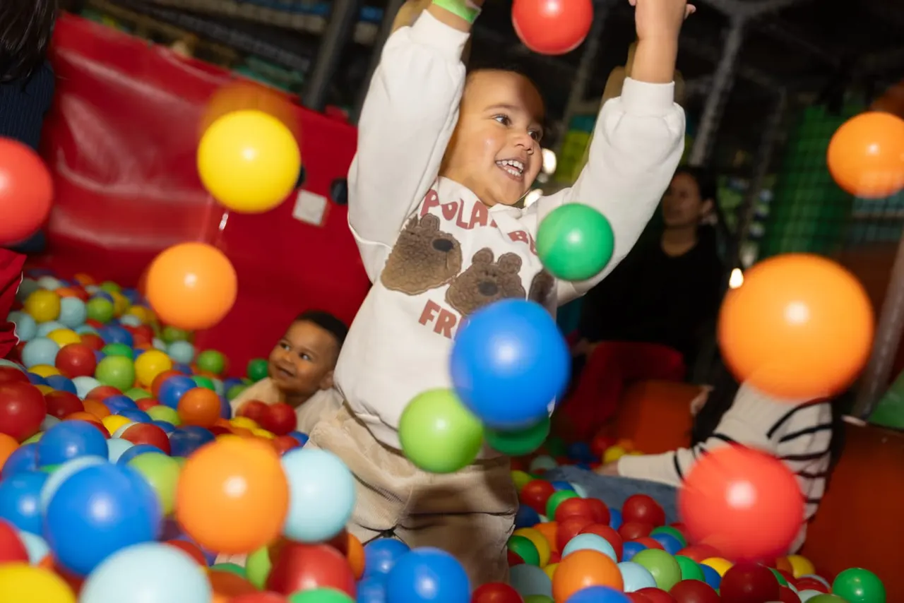 Children playing in a colourful ball pit, throwing and catching plastic balls in an indoor play area.