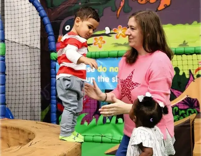 An adult supervises two young children in a colourful indoor trampoline park with padded walls and safety netting.