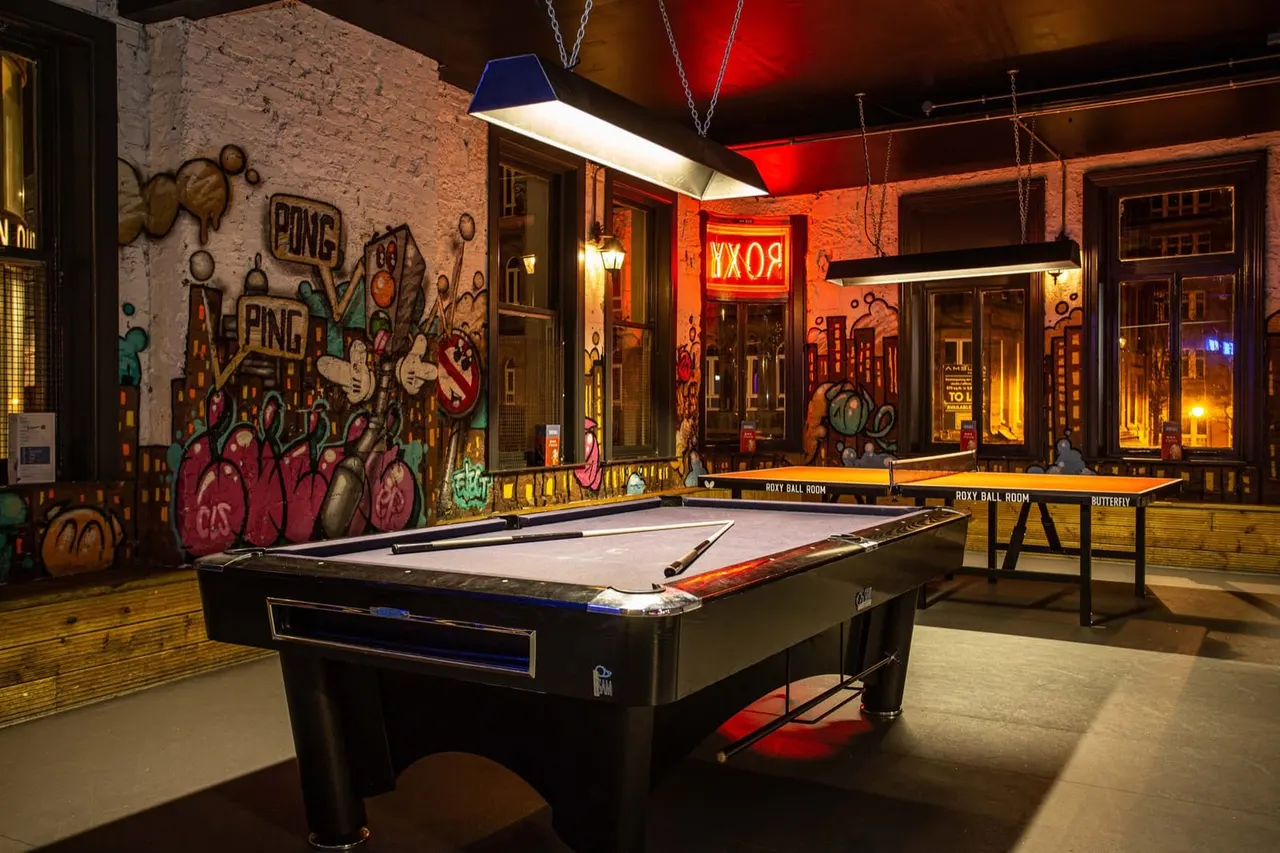 Pool tables in an indoor games room with colourful street art murals on brick walls and neon signage.