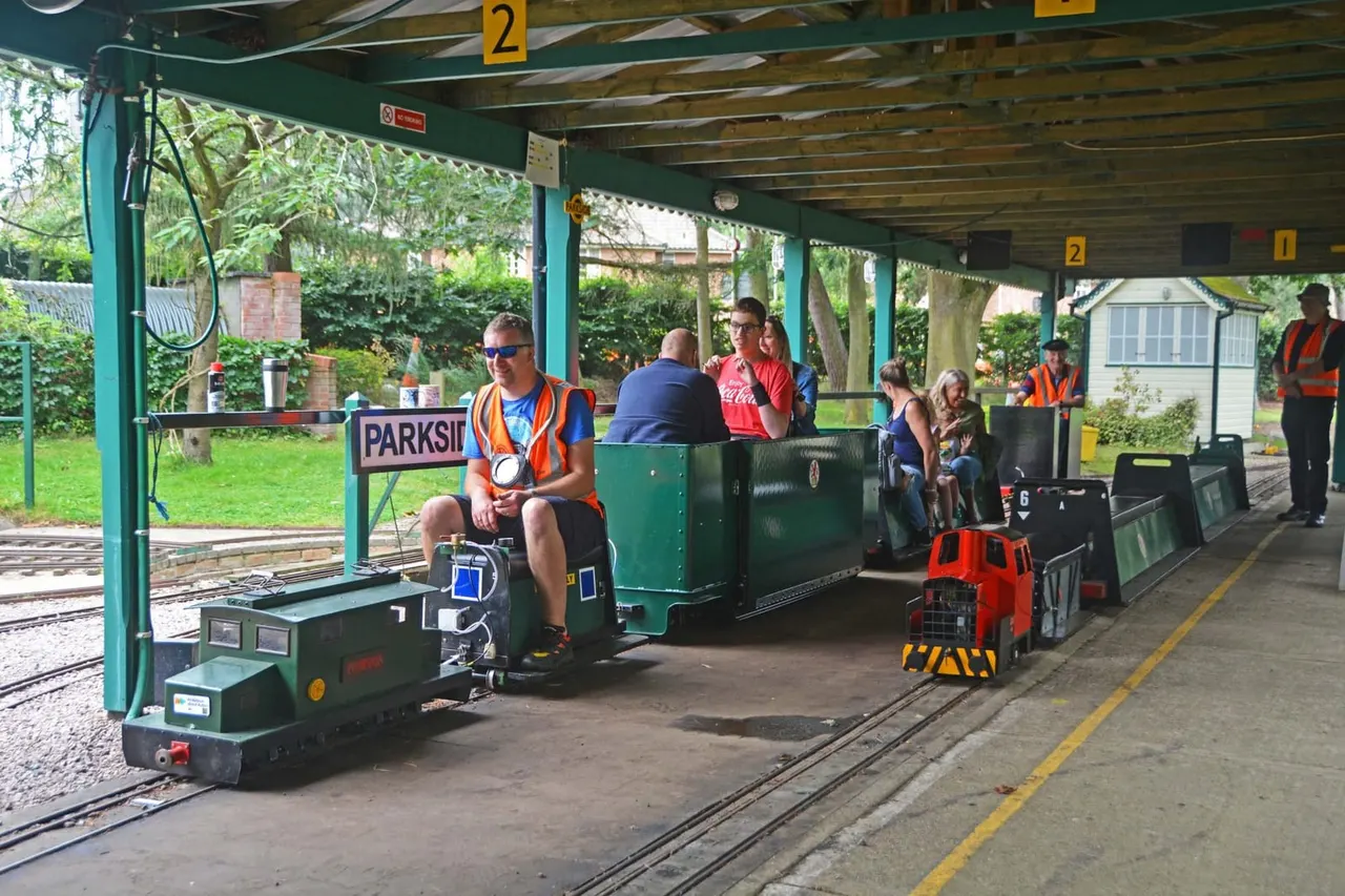 Passengers board a miniature railway train at a covered station platform surrounded by green landscaping.