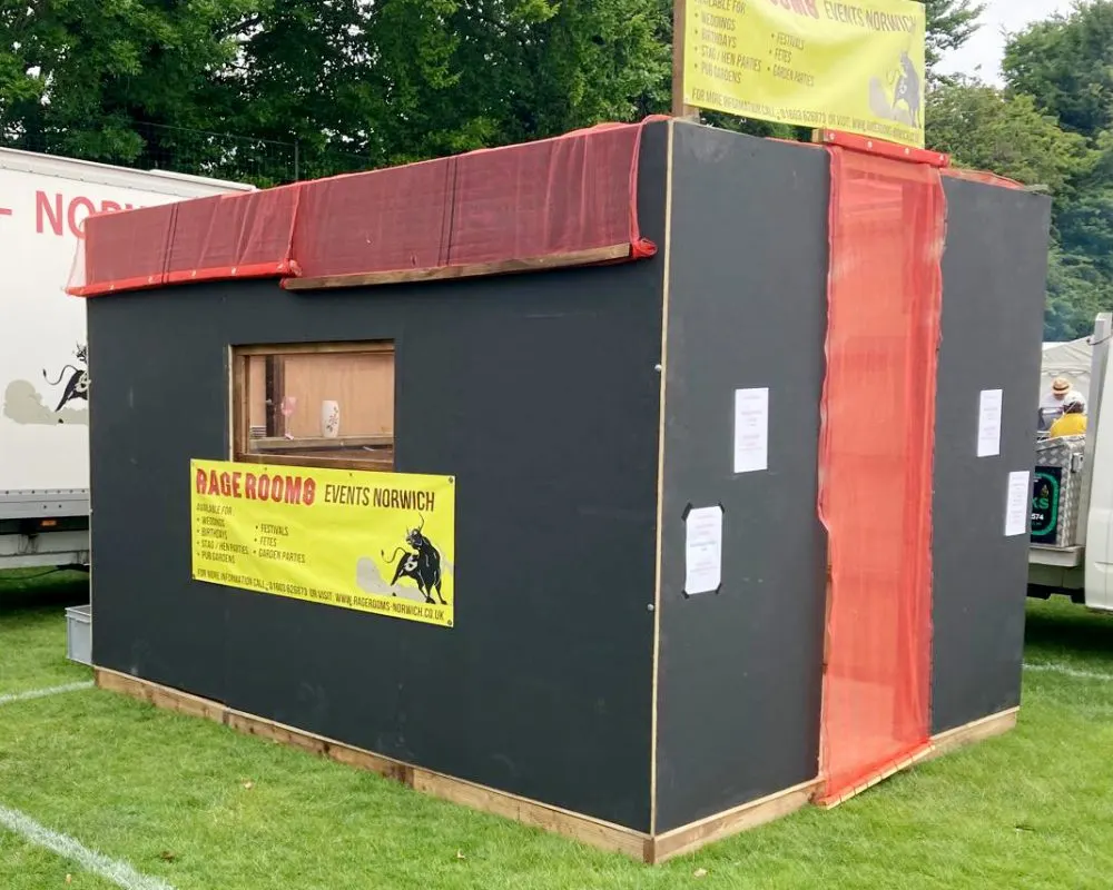 A portable black and red rage room booth on grass with yellow promotional signage and trees in the background.