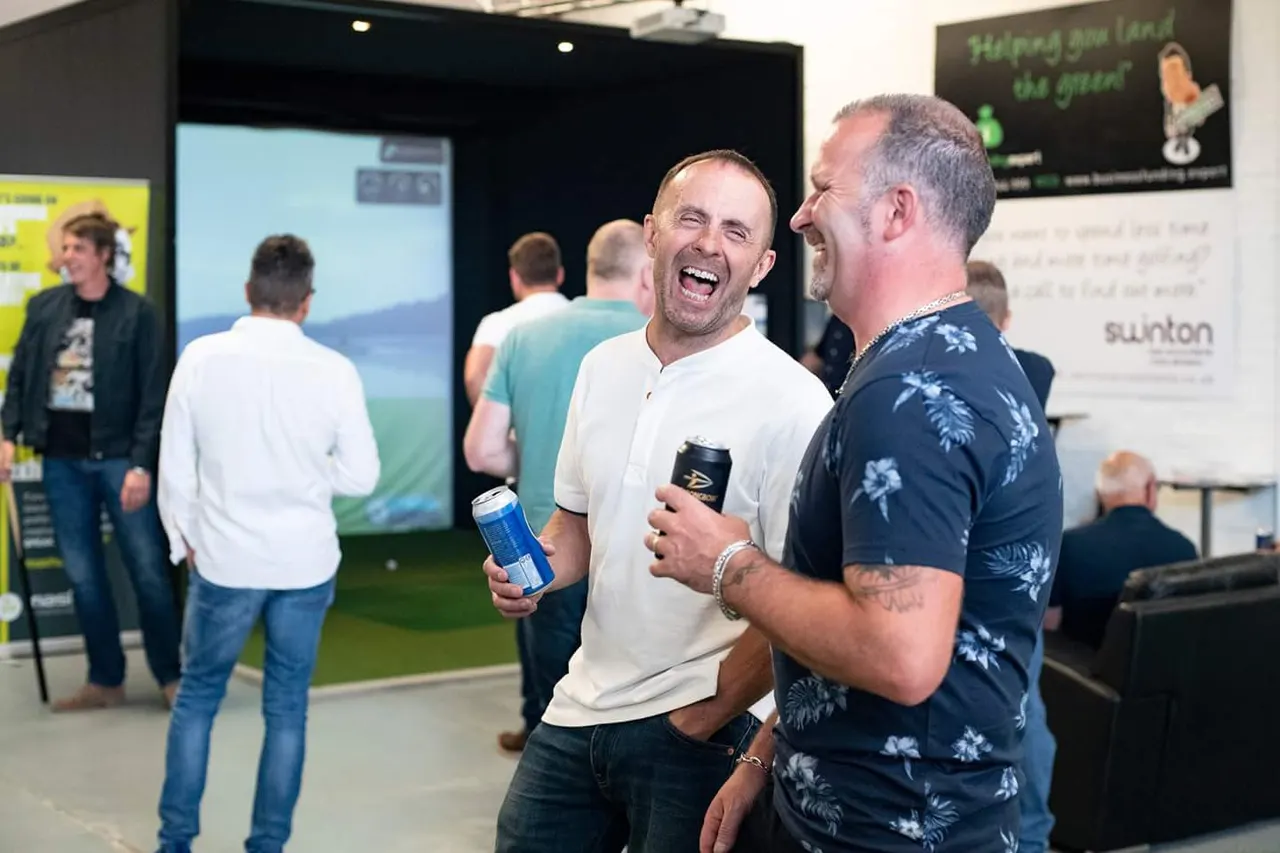 Two men laughing whilst holding drinks at an indoor social gathering, with other guests mingling in the background.