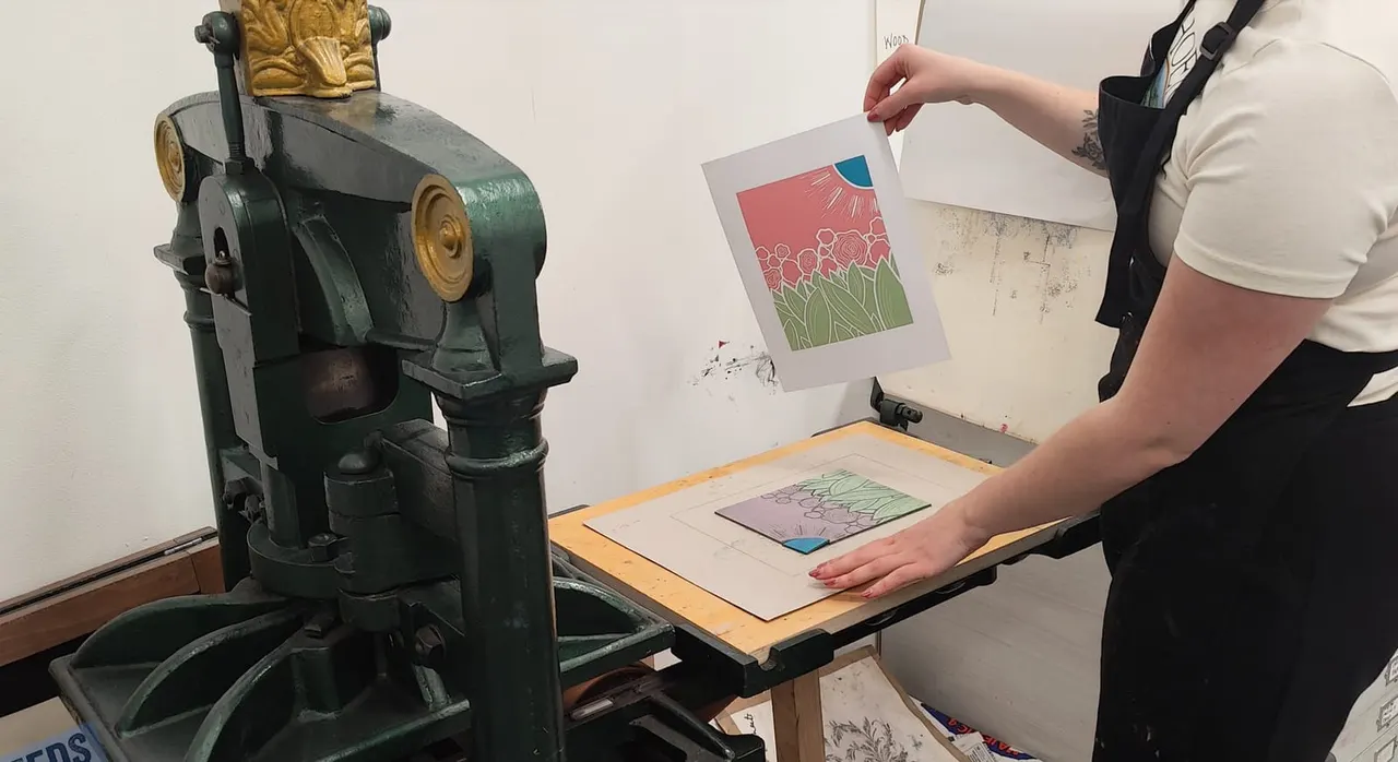 Person demonstrating a traditional printing press, holding a printed sheet with floral artwork design.