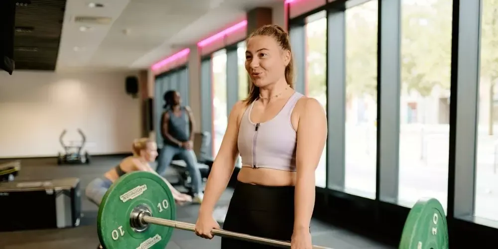 Woman holding a dumbbell in a gym with pink neon lighting and large windows, other gym users visible in the background.