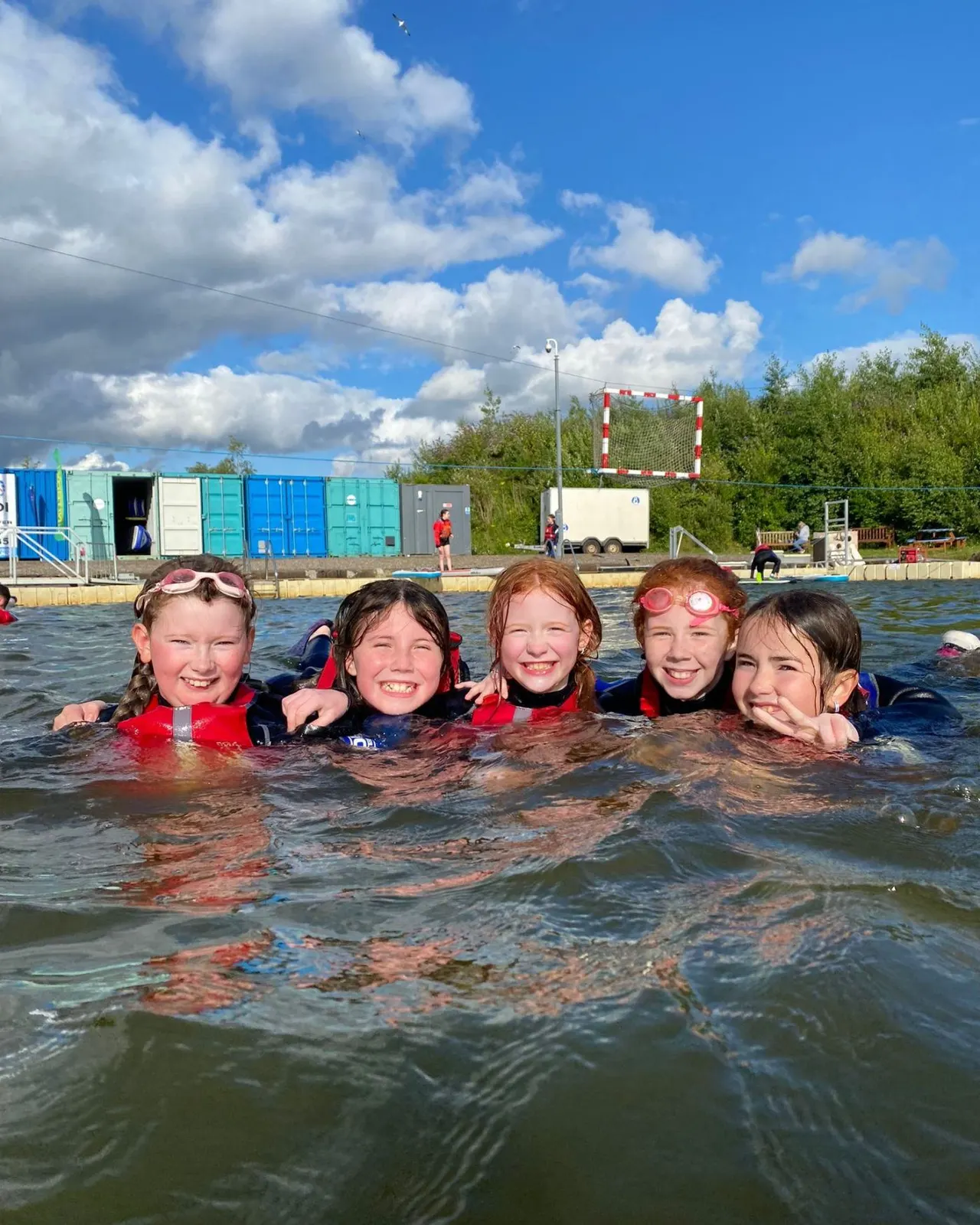 Five children in swimming costumes and buoyancy aids smile at the camera whilst in the water at an outdoor watersports facility.