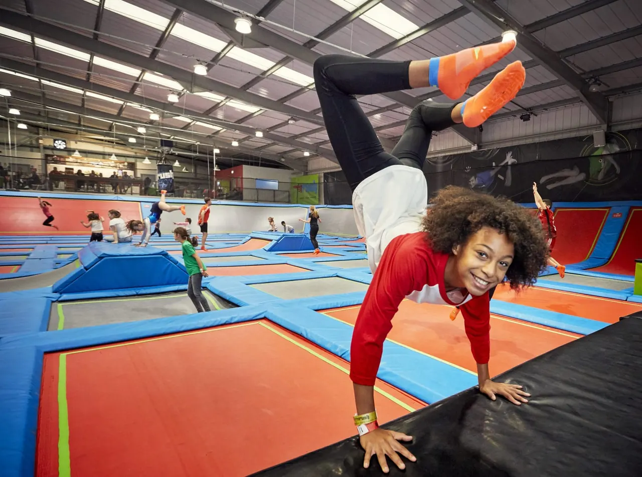 Young person doing a backflip on a coloured trampoline whilst others jump and play in an indoor soft-play facility.