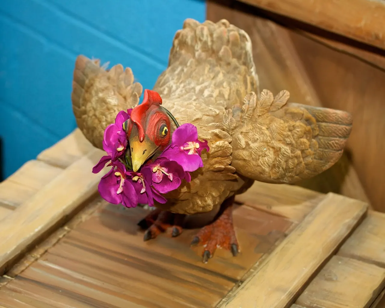 Decorative wooden bird sculpture holding bright magenta flowers, displayed on a wooden deck beside a blue water feature.