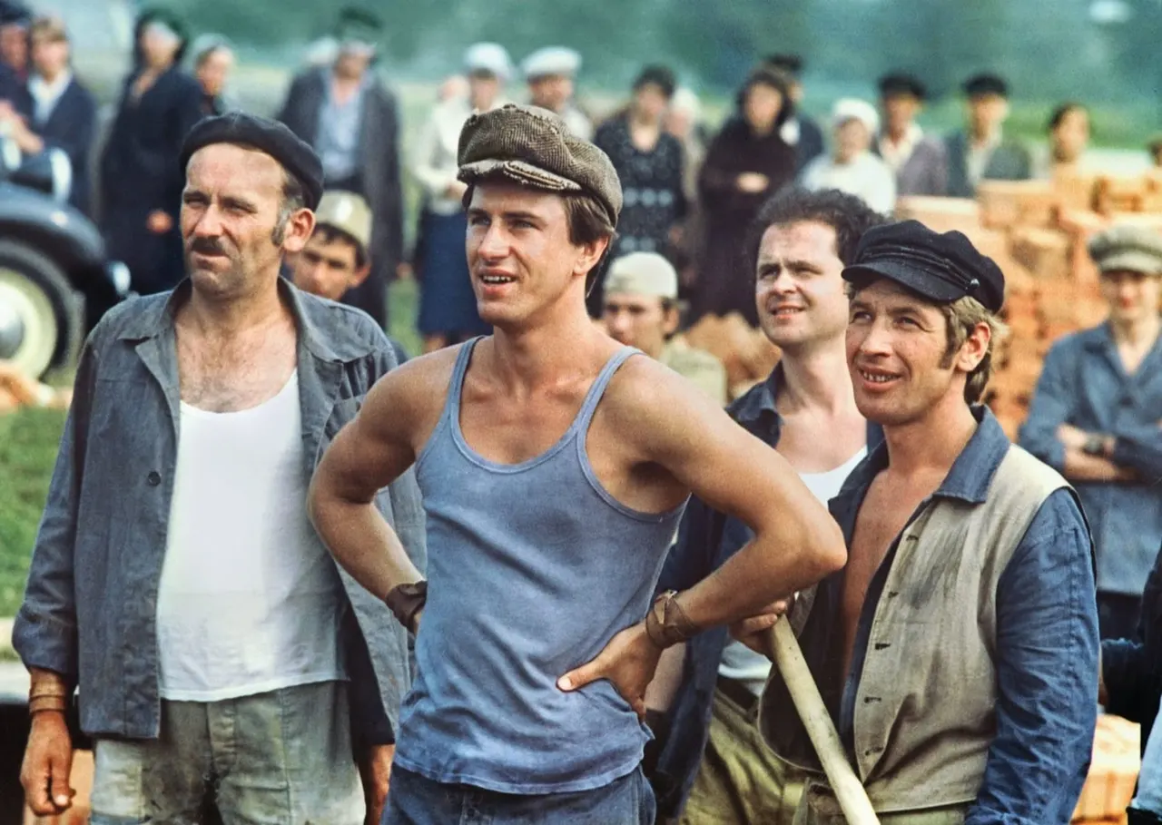 A group of men in casual work clothes stand together outdoors whilst a crowd watches in the background.