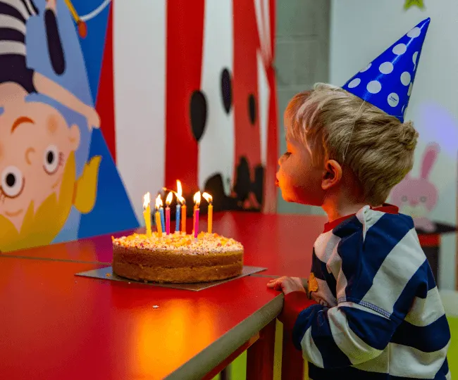 Young child in blue party hat gazing at lit birthday cake on red table with colourful decorations.