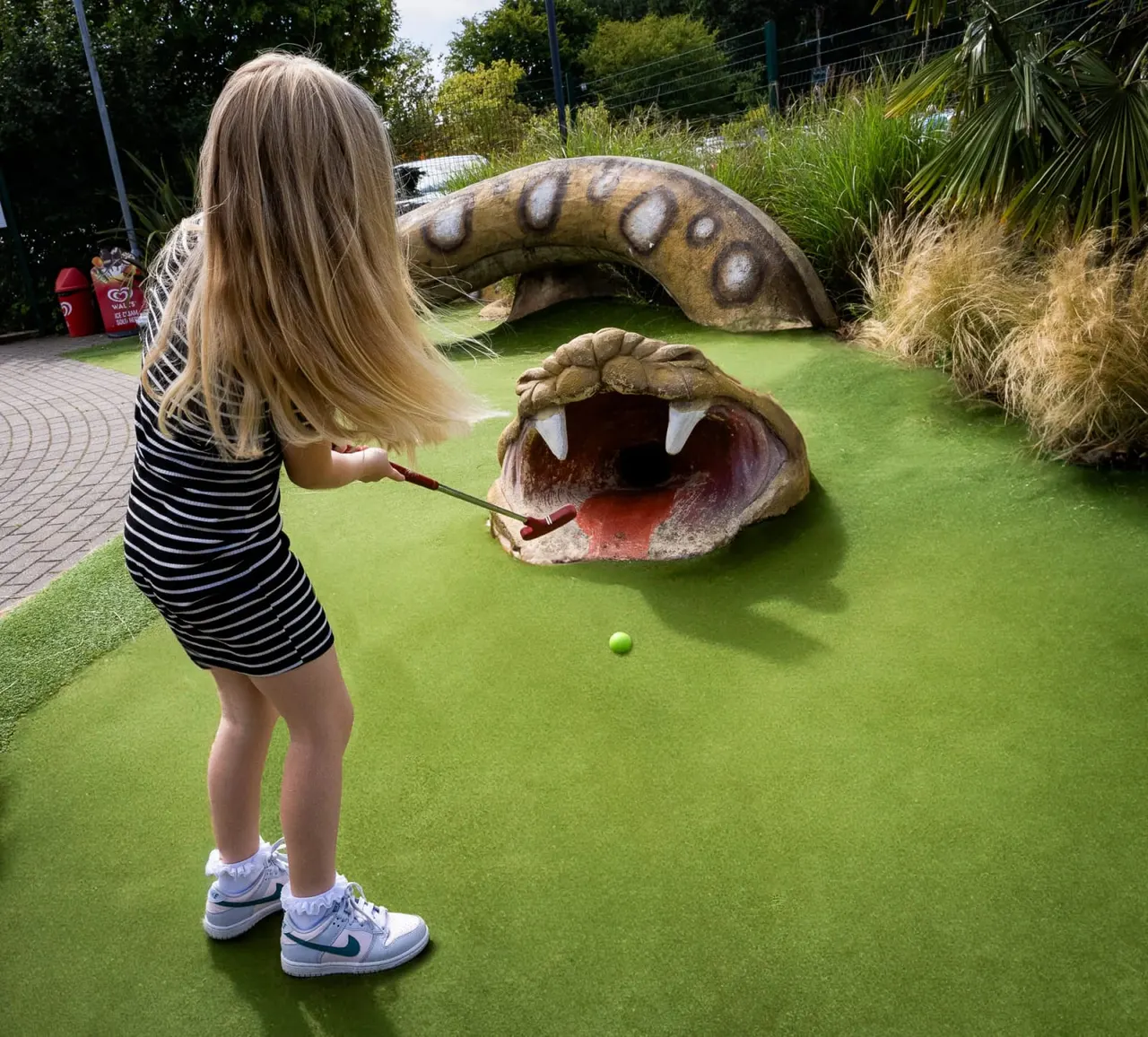 Child playing miniature golf, putting towards a large themed crocodile obstacle with open jaws.
