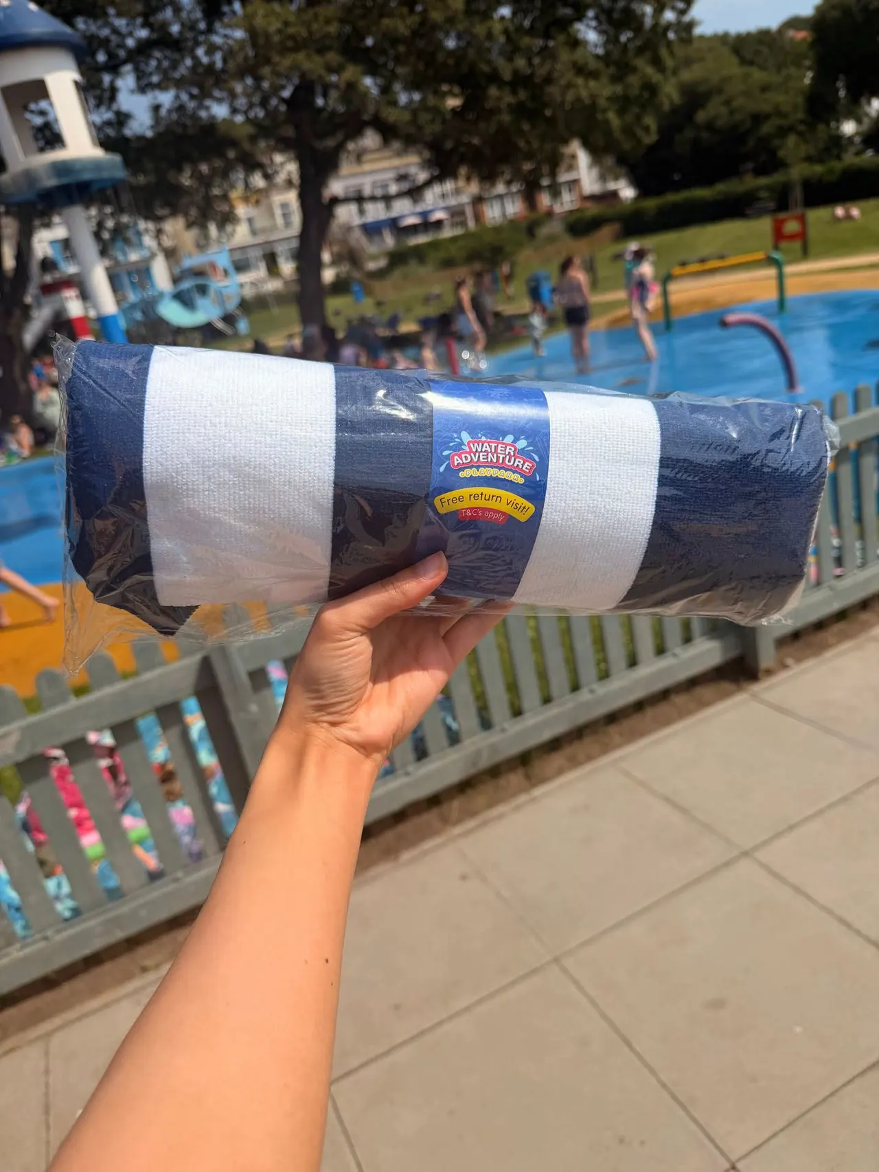 Hand holding a water adventure souvenir tube with visitors playing in the paddling pool behind.