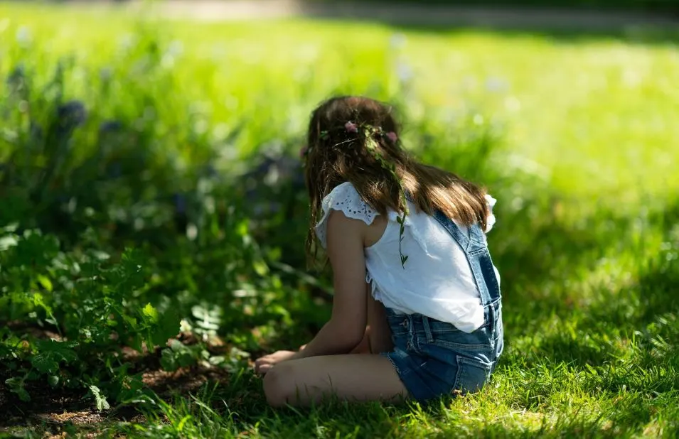 Child crouching on grass in sunlit garden, picking flowers.