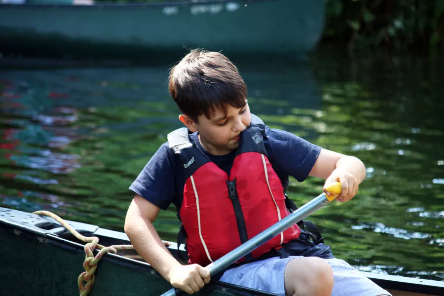 Young child wearing a red life jacket paddling a small boat on calm water.