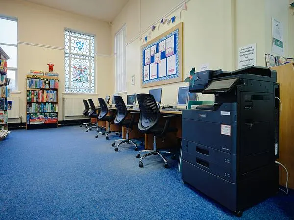 Library computer room with several desktop workstations, a photocopier, bookshelf and notice boards on cream walls.
