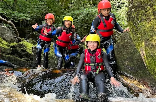 Four children in safety helmets and life jackets sit on a rock in a fast-flowing stream surrounded by woodland.