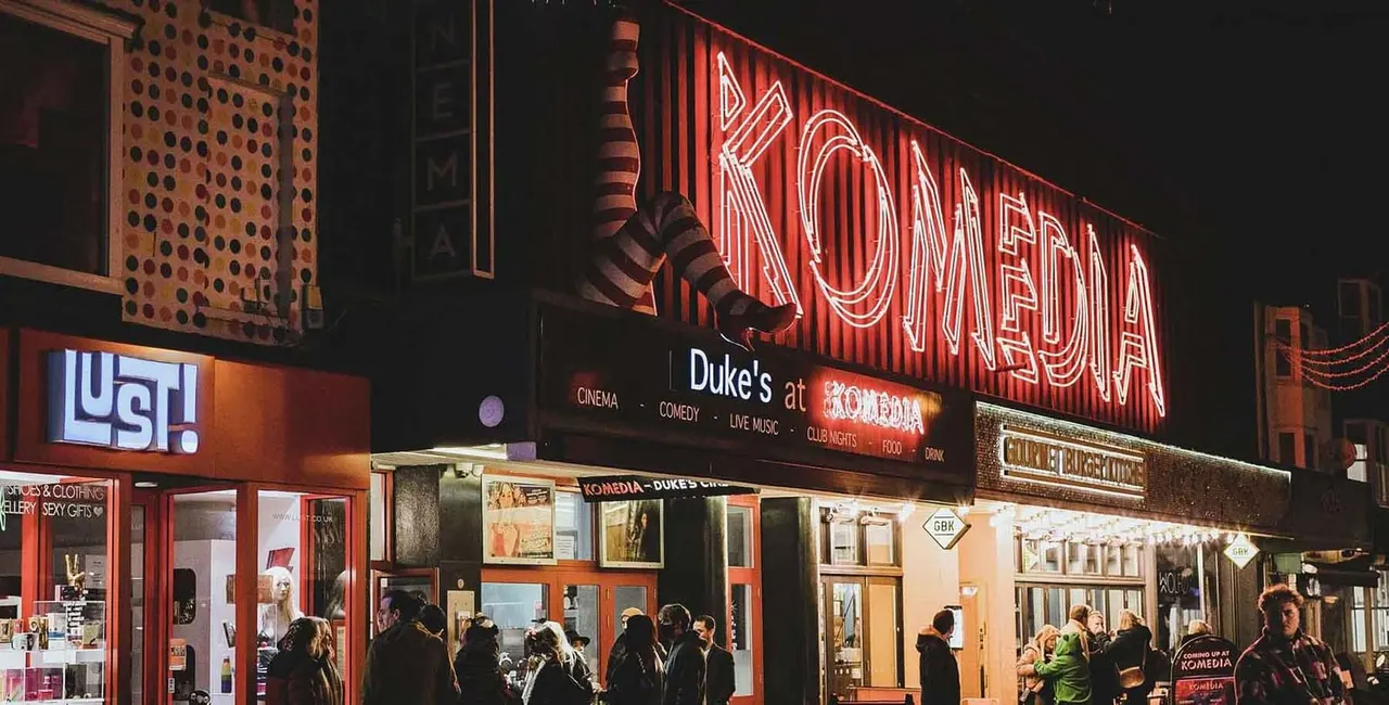 A nighttime street scene showing the illuminated red neon sign for Komedia entertainment venue, with people gathered outside the entrance.