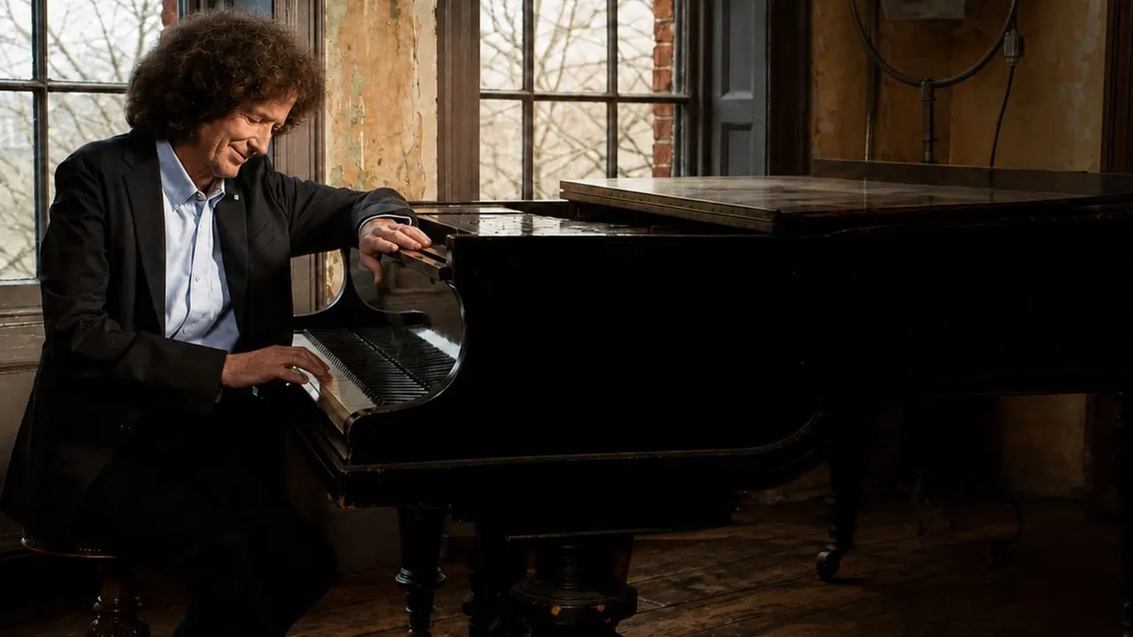 Man in a black jacket playing a grand piano in a room with large windows and exposed brick walls.