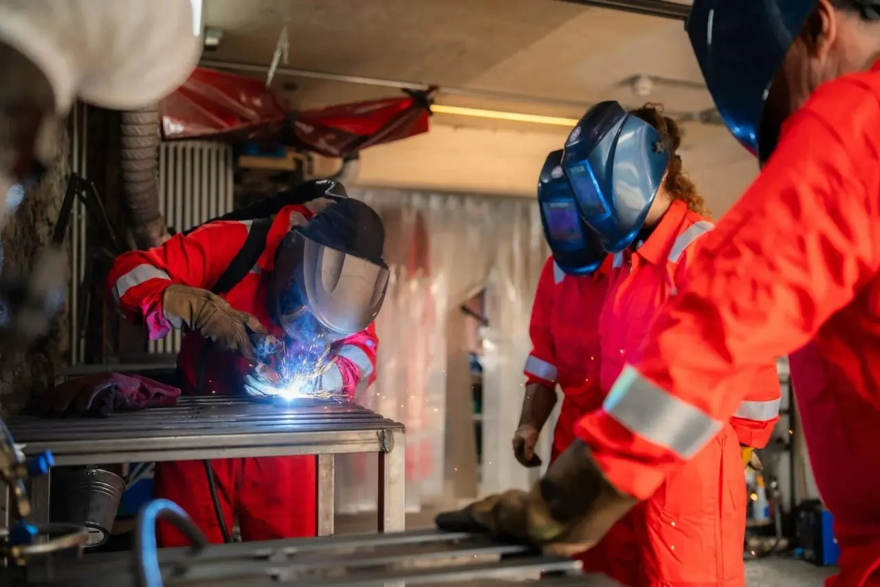 People in red protective suits and welding masks practise welding metal indoors.