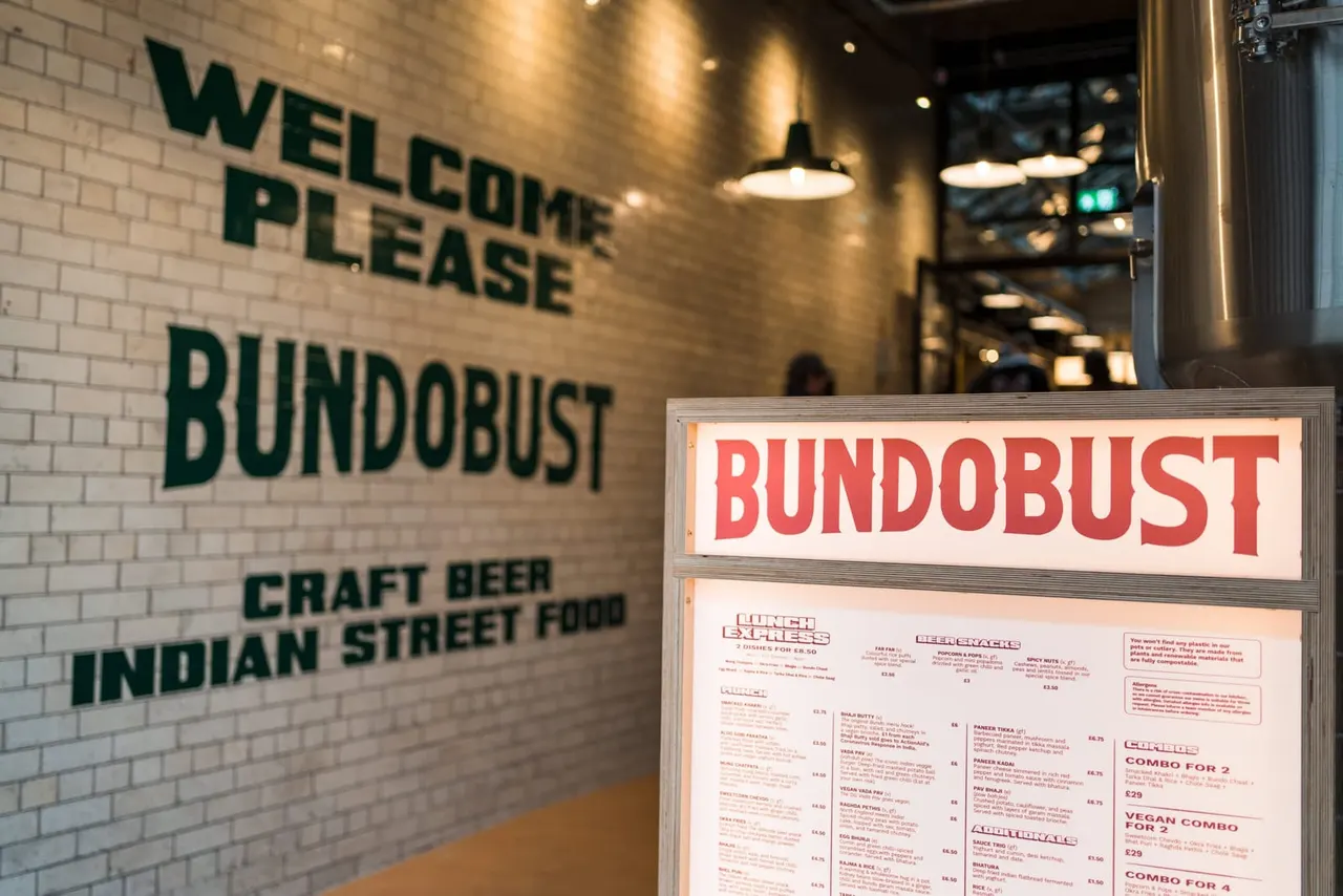 Interior of a casual dining venue with a white brick wall displaying a welcome sign and menu board, featuring pendant lighting and an illuminated red "Bundobust" sign.